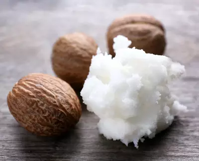 Alt text: Four whole nutmegs and a scoop of white, soft shea butter on a wooden surface.