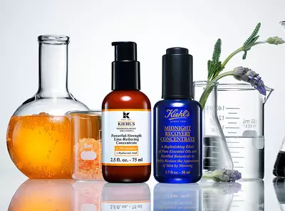Ket Kiehl's bőrapolo termek uvegben, az egyik narancssarga, a masik kek, gyogynovenyek es uvegek kozott.
