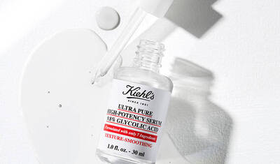 A bottle of Kiehl's Ultra Pure High-Potency Serum containing 38% glycolic acid, accompanied by a droplet of the serum on a white surface. The label emphasizes texture-smoothing benefits.