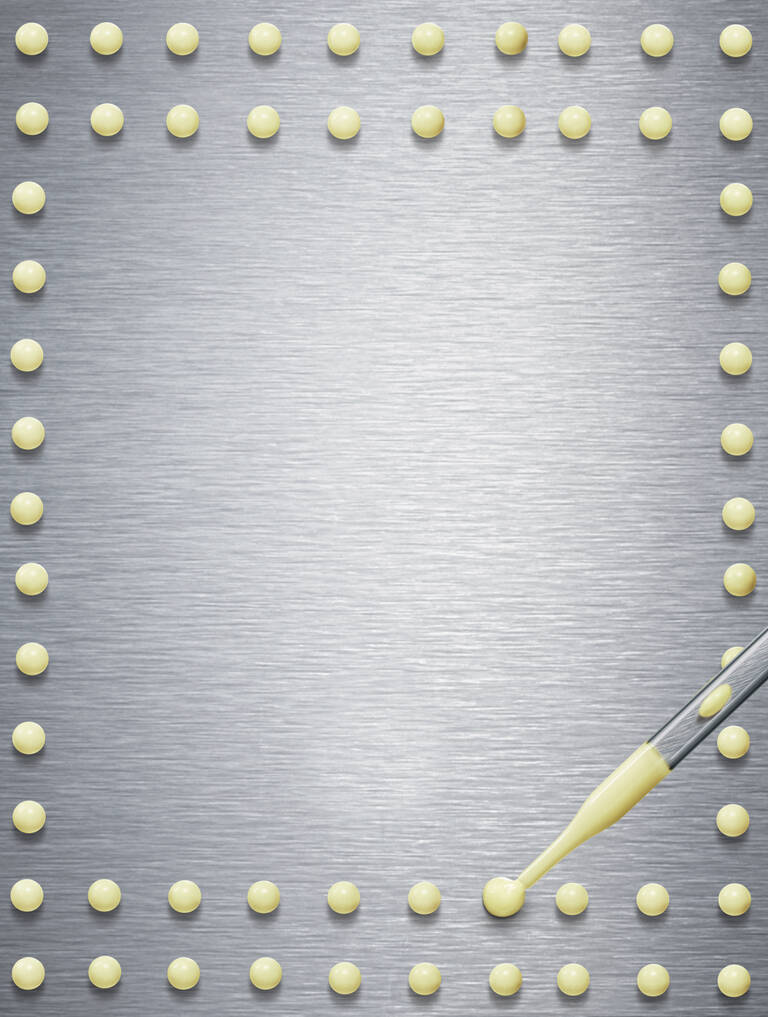 A close-up of a metallic surface with evenly spaced small yellow dots arranged in a pattern, and a pipette poised above one of the dots, ready to dispense a drop of liquid.