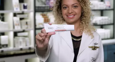 A smiling woman with curly hair holds a tube of Kiehl's cream in a retail setting, surrounded by skincare products on shelves.