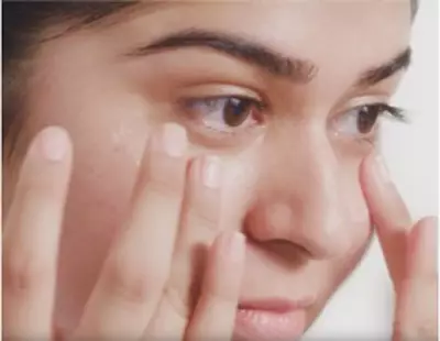A close-up of a woman applying cream to her face, focusing on her under-eye area, with her fingers gently massaging the product in.