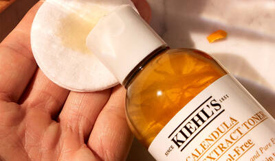 A hand holding a cotton pad with liquid being dispensed from a bottle of Kiehl's Calendula Extract Toner.