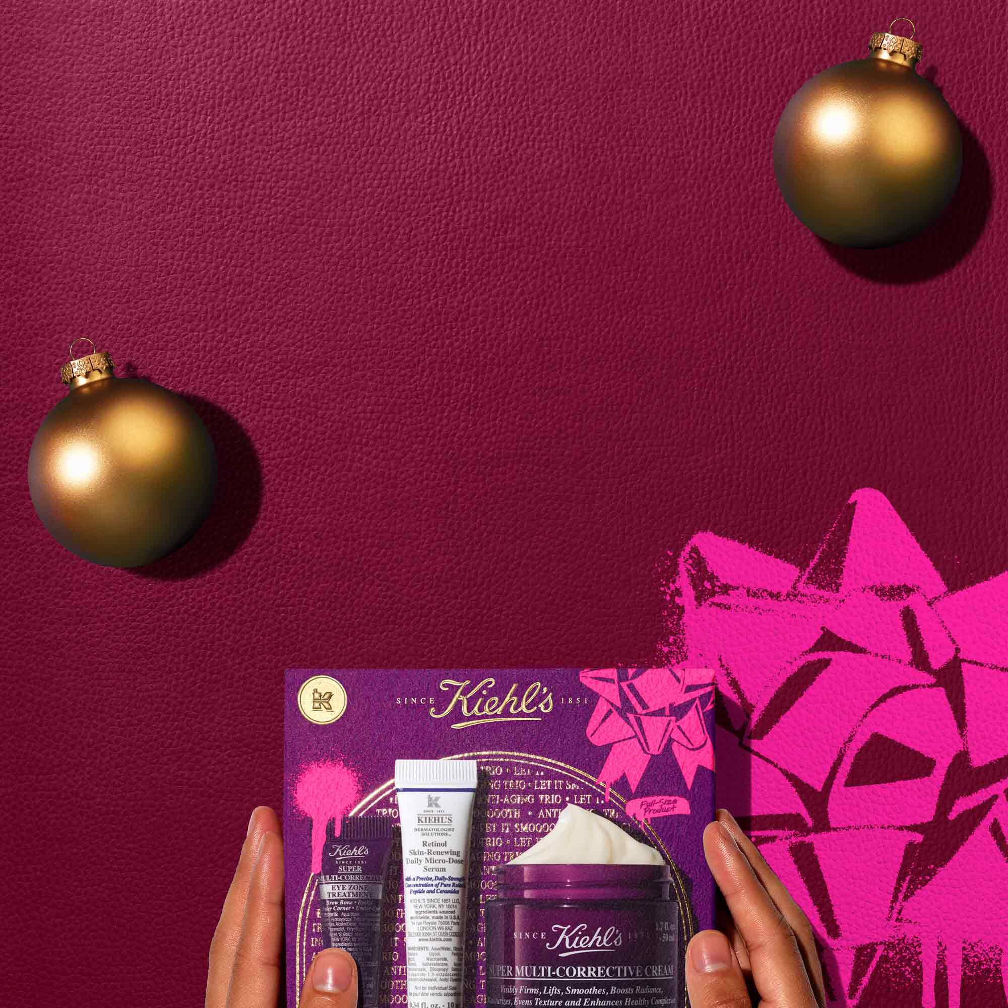 A graphic, set against a dark red textured background, features three Kiehl’s products and a gift box. The top row displays a small purple tube of "Kiehl's Super Multi-Corrective Eye Zone Treatment," a white and purple tube of "Kiehl's Retinol Skin-Renewing Daily Micro-Dose Serum," and an open purple jar of "Kiehl's Super Multi-Corrective Cream" with visible cream. Two gold spherical ornaments are also present. Below these, a purple Kiehl’s gift box showcases the same three products, decorated with bright pink graphic paint splatters and a bow design.