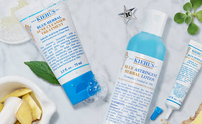 A flat lay image of Kiehl's skincare products, including a blue herbal acne treatment, a blue astringent herbal lotion, and a small herbal treatment tube, surrounded by fresh herbs and natural ingredients on a marble surface.