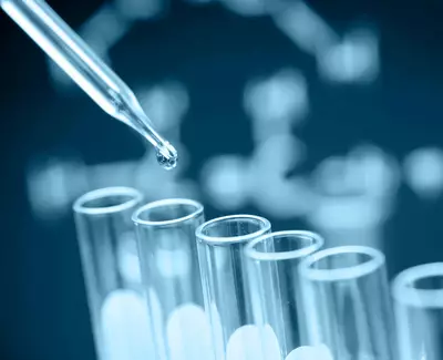 A close-up of a pipette dropping a liquid into a set of test tubes, with a blurred background featuring a DNA structure.