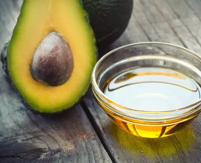 A sliced avocado revealing its seed next to a small glass bowl of golden avocado oil, placed on a wooden surface.