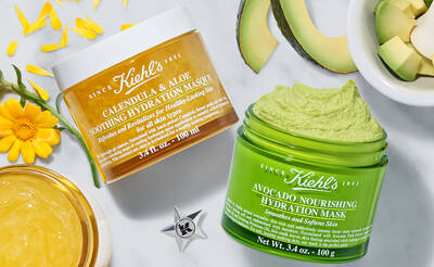 Two jars of Kiehl's skincare masks are displayed on a textured surface. The first, labeled "Calendula & Aloe Soothing Hydration Mask," is a cream-colored jar; the second, "Avocado Nourishing Hydration Mask," is a green jar. Surrounding the jars are petals, avocado slices, and a small bowl of gel