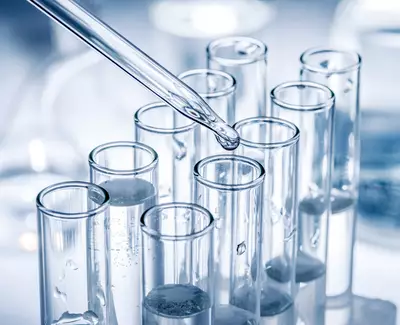A close-up of a glass test tube rack filled with clear liquid, with a dropper positioned above one of the tubes, poised to dispense a drop.