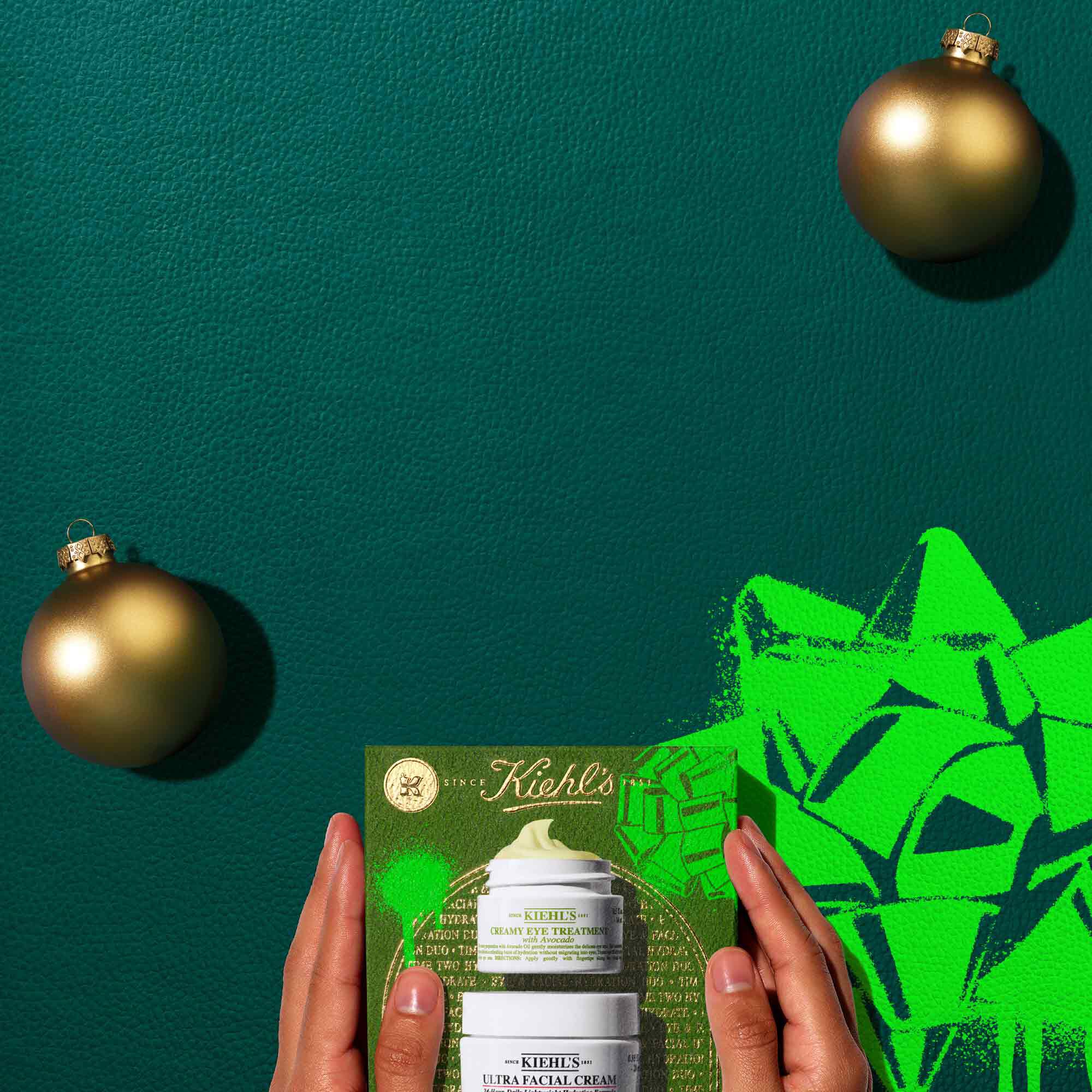 A graphic of an open green Kiehl's gift box, held by two hands, revealing its contents. There is a bright green graphic bow in the background.. Inside the box are two Kiehl's jars: "KIEHL'S CREAMY EYE TREATMENT with avocado" showing a yellow-green product swirl, and "KIEHL'S ULTRA FACIAL CREAM". Two gold Christmas ornaments are also part of the scene. All set against a dark green background.
