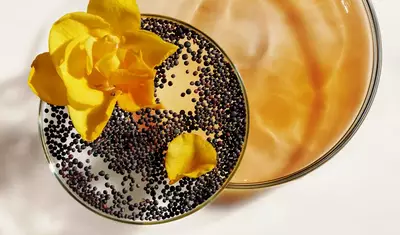 A close-up of two glass bowls, one filled with a light brown liquid and the other containing black seeds, adorned with a yellow flower petal.
